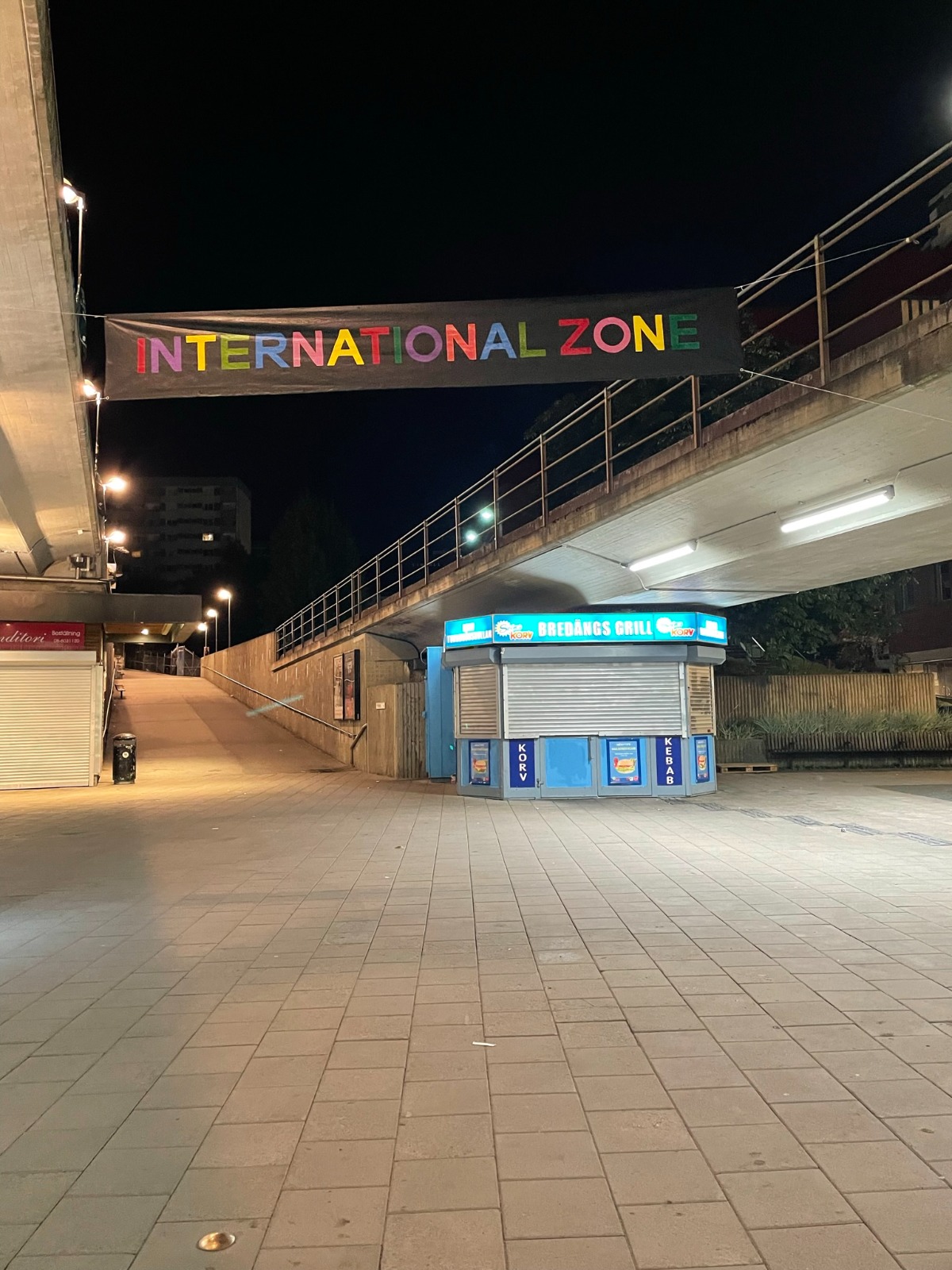 INTERNATIONAL ZONE-bannern hänger upplyst i kvällsmörker över Bredängs torg.