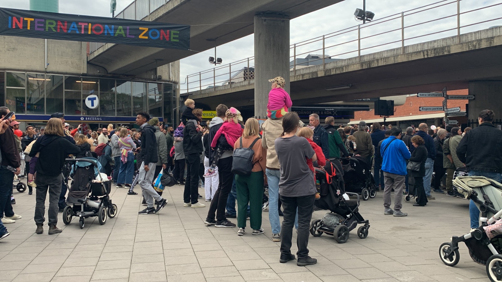 Folkmassa vid Bredängs tunnelbanestation under den färgglada bannern.